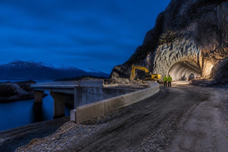 Veianlegg med bru langs fjorden og tunnelåpning der to arbeidere står ved en gravemaskin