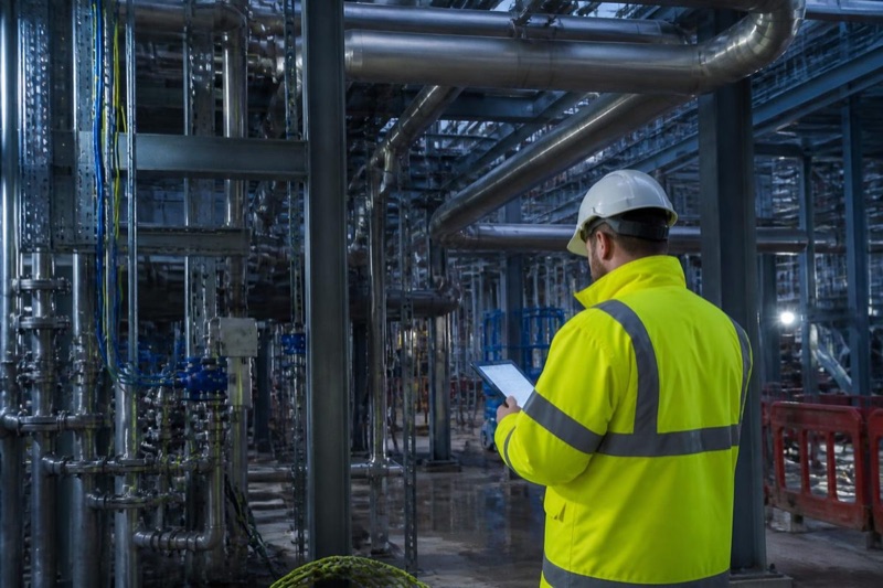 Arbeider med hjelm og nettbrett i et industrianlegg med rør og stålkonstruksjoner