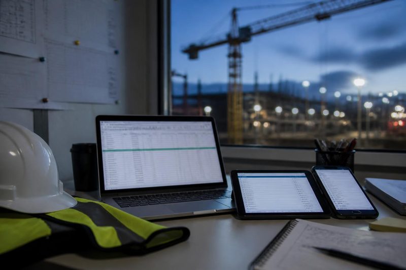 Bærbar PC, nettbrett og mobil med regneark på et bord i en anleggsbrakke, med kran og byggeplass utenfor vinduet